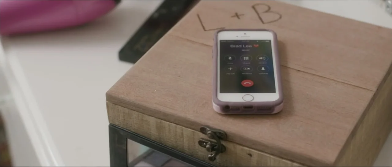 If You're Gone (2019) movie still: closeup — A wooden jewelry box sits on a white surface with a smartphone placed atop it. The phone screen shows…