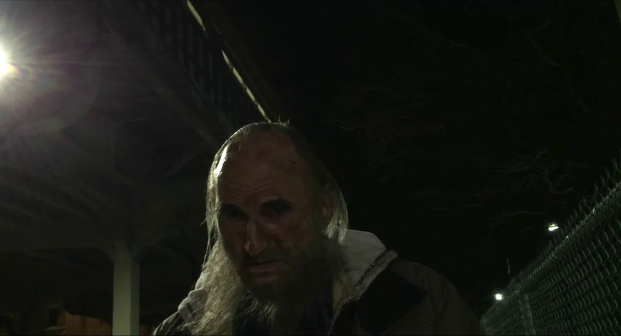 Like Me movie still: closeup — An outdoor area at night with a chain-link fence and a dark structure overhead. An older man with a long beard stands…