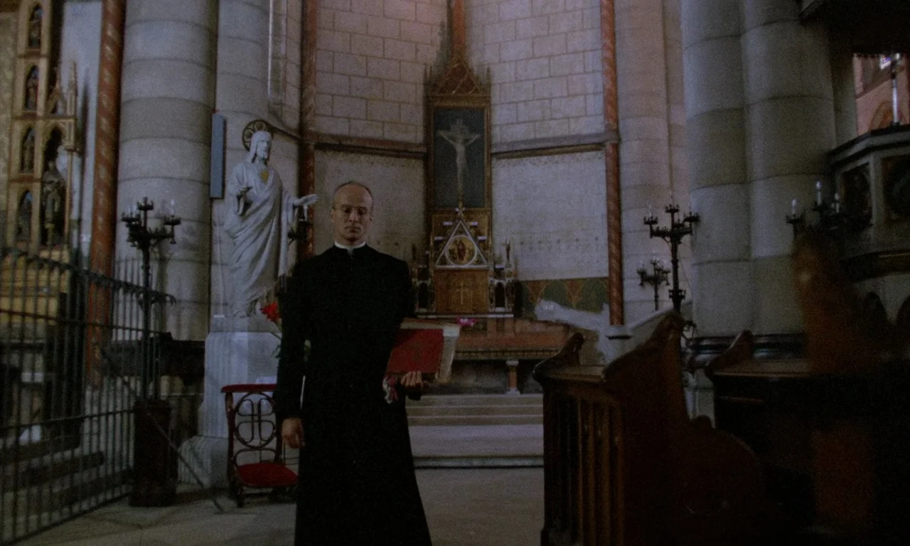 The Church (1989) movie still: fullbody — A stone cathedral nave features large columns and a central altar. A priest in a black cassock stands…