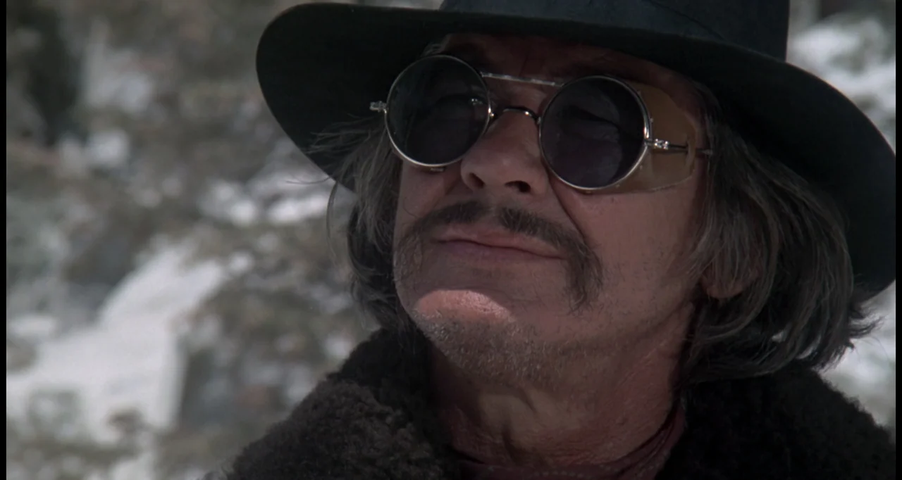The White Buffalo (1977) movie still: closeup — A man stands outdoors in a mountainous region during the day. He wears a wide brimmed hat and round…