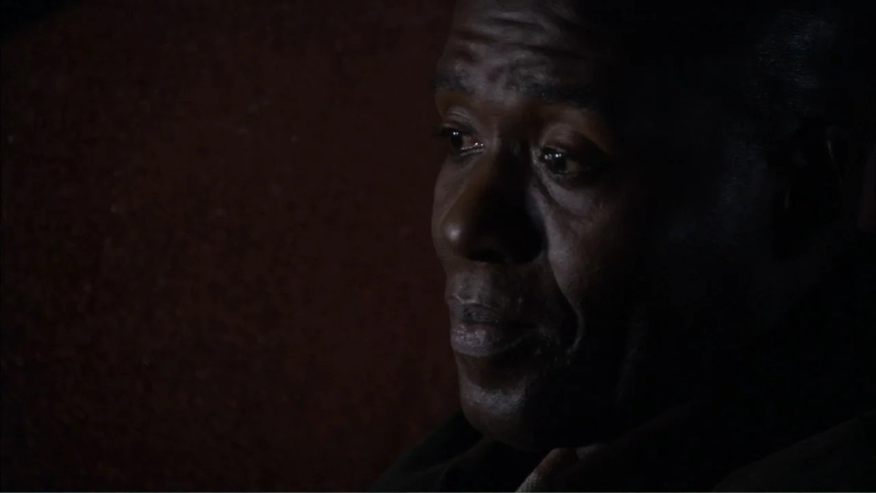 O código oculto (2025) movie still: closeup — A dark, nondescript interior space with a textured wall visible in the background. An older Black man…