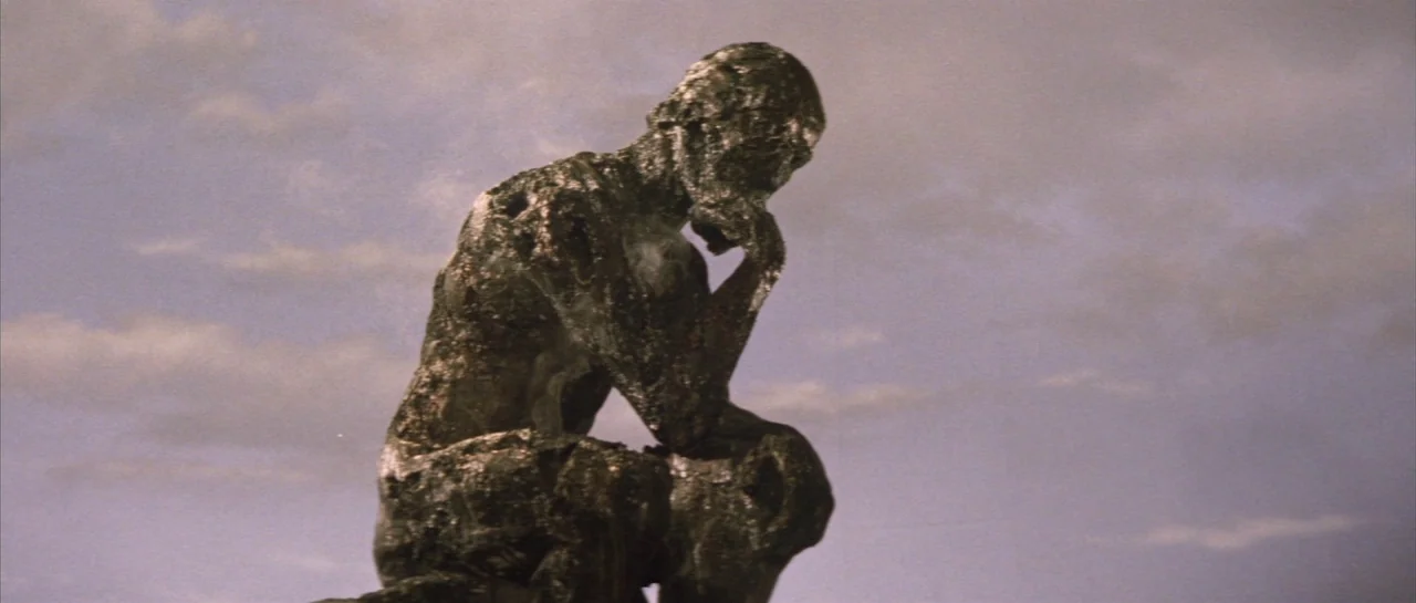 Godzilla Vs Hedorah (1971) movie still: medium — A bronze cast of The Thinker statue stands against a pale, overcast sky. The dark metallic…