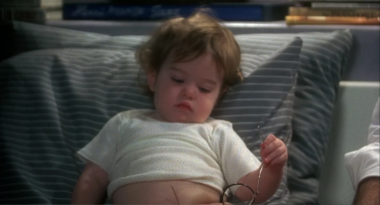 Baby Boom (1987) movie still: medium — A toddler rests against grey textured pillows inside a domestic living space. She holds a pair of eyeglasses…