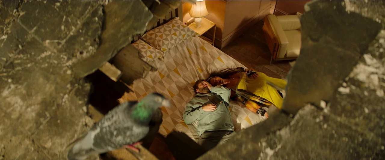Unwelcome (2022) movie still: wide — A damaged room shows a gaping hole in the wall above a double bed. A man and woman lie side by side on the bed…