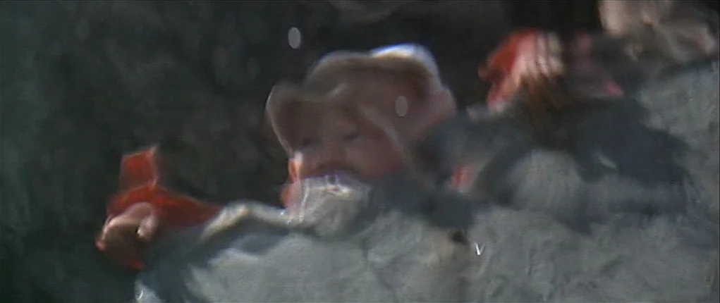 Popeye (1980) movie still: closeup — A child in light-colored headwear appears in motion against a textured, indistinct background. The subject is…