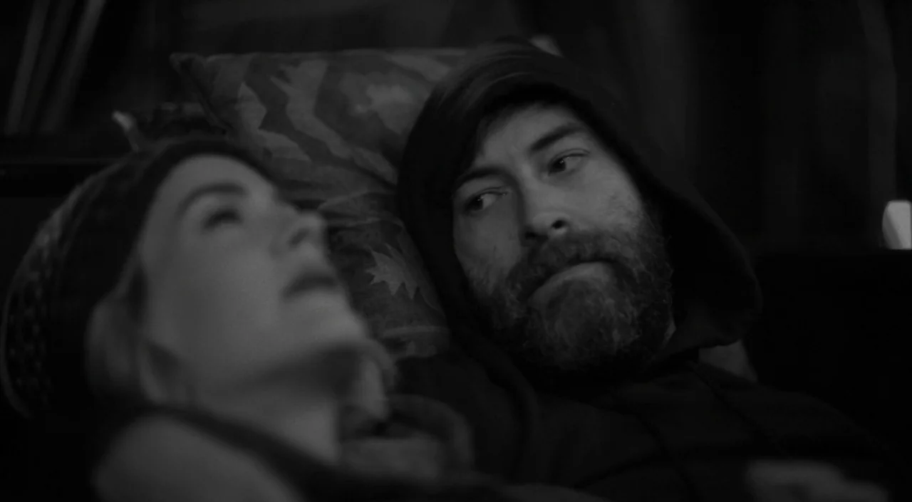 Blue Jay (2016) movie still: closeup — A dimly lit interior scene features a couple resting close together on a couch. The man wears a hood while…