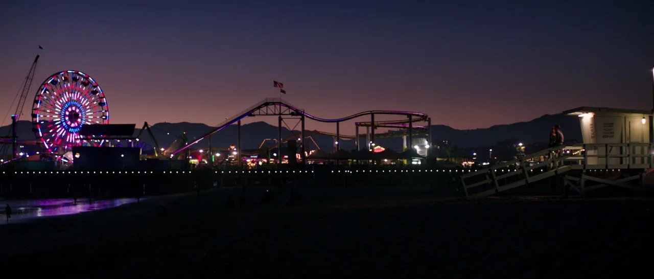 Wish I Was Here (2014) movie still: wide — A seaside pier features a illuminated ferris wheel and roller coaster at night. The dark beach is in the…