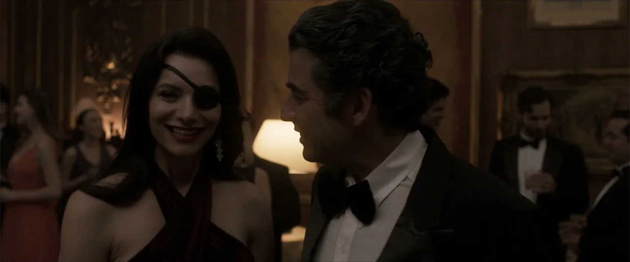 Angela (2023) movie still: medium — A dimly lit room with ornate walls serves as a backdrop for a gathering. A woman wearing an eye patch and a man…