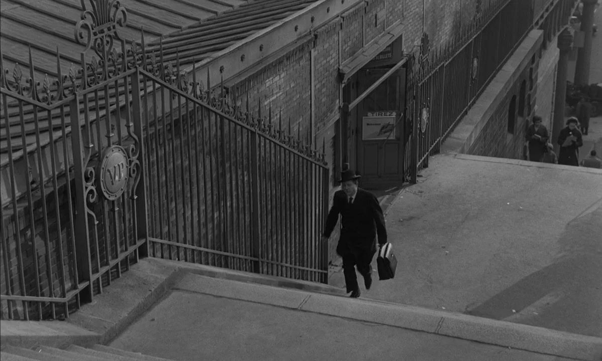 La Peau Douce (The Soft Skin) (1964) movie still: wide — An outdoor stone staircase runs alongside a long brick wall with an ornate gate. A man in a…