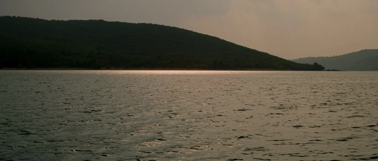 Stranger By The Lake (2013) movie still: establishing — A dark, hilly shoreline borders a calm expanse of water under an overcast sky. The sun…