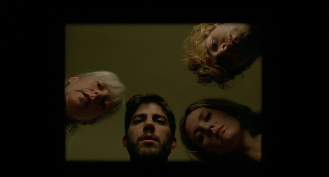 Tom at the Farm (2013) movie still: medium — Four people gather around looking down from above against a flat, neutral-colored background. They gaze…