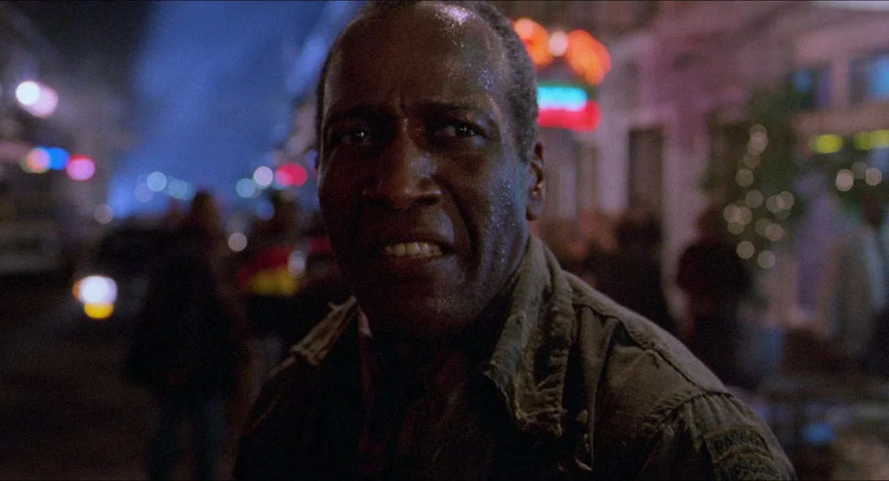 Hard Target (1993) movie still: closeup — A nighttime city street illuminated by colorful blurred neon signage and urban light sources. A…