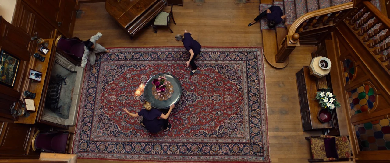 Polite Society (2023) movie still: wide — A spacious home interior featuring dark polished wooden panels and an ornate patterned carpet. Four people…