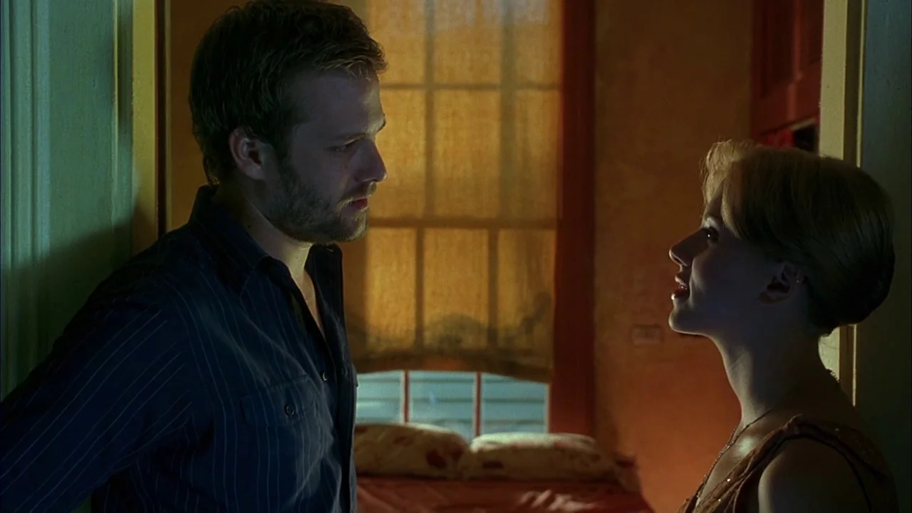Love Song For Bobby Long (2004) movie still: medium — An interior shot shows two people standing in a dimly lit, amber-toned space. The man and…