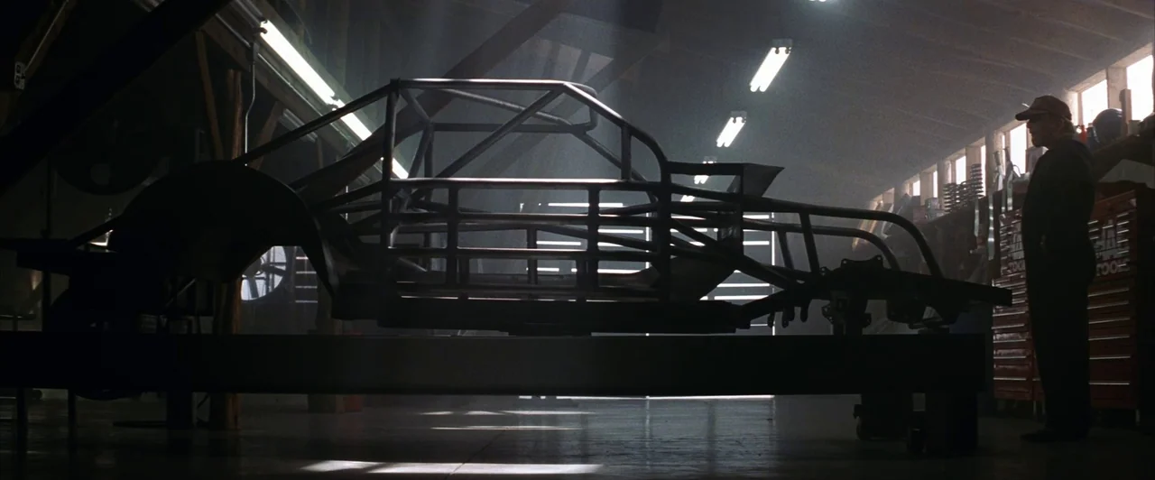 Days of Thunder (1990) movie still: wide — A dark auto repair shop contains a prominent steel race car chassis in the center. An older man stands to…