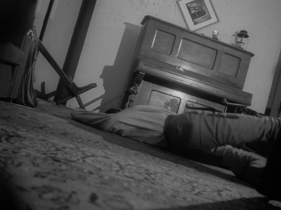 Night of the Living Dead (1968) movie still: fullbody — A domestic space features a wooden upright piano against a plain wall. A man lies motionless…