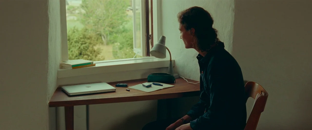 Bergman Island (2021) movie still: medium — A workspace features a minimalist wooden desk placed in front of an open window. A woman sits at the…