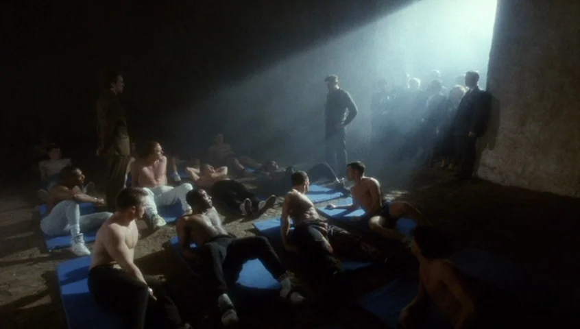 Edward II (1991) movie still: wide — A dimly lit industrial room features a dusty stone wall and scattered blue exercise mats. A group of shirtless…