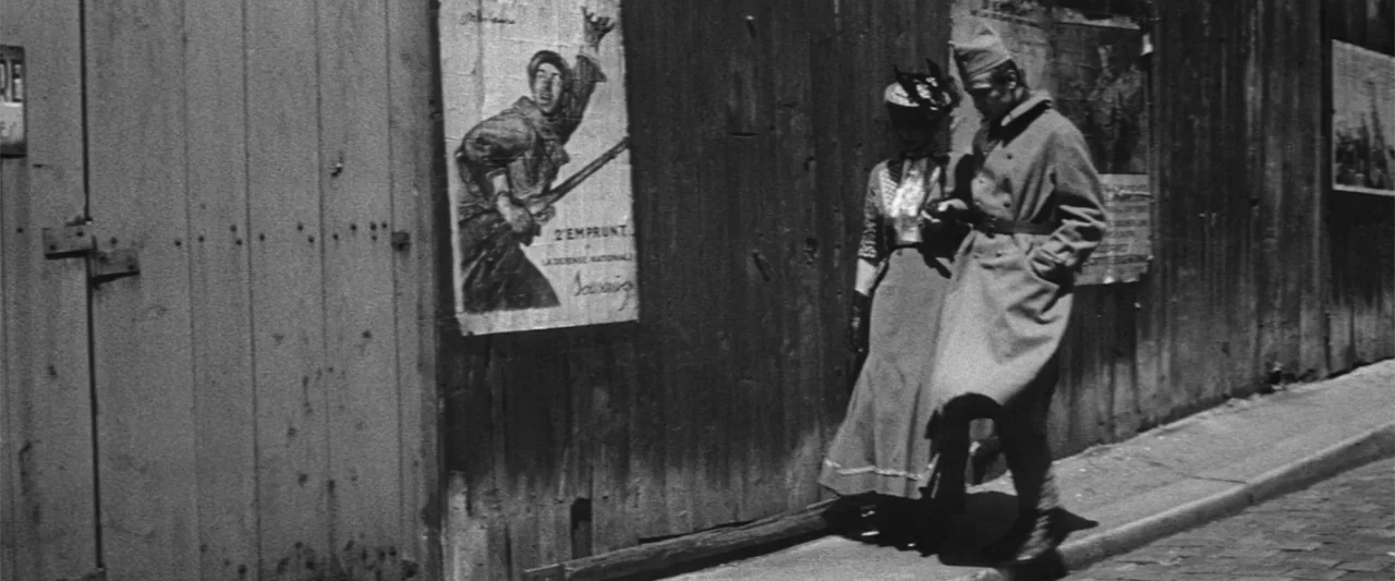 Jules and Jim (1962) movie still: fullbody — A street with a large vertical wooden plank wall displaying vintage French war posters. A man in a…
