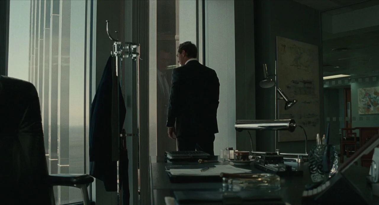 The Nest (2020) movie still: wide — An upscale professional office space with dark wood furniture and high-rise glass walls. A man in a suit stands…