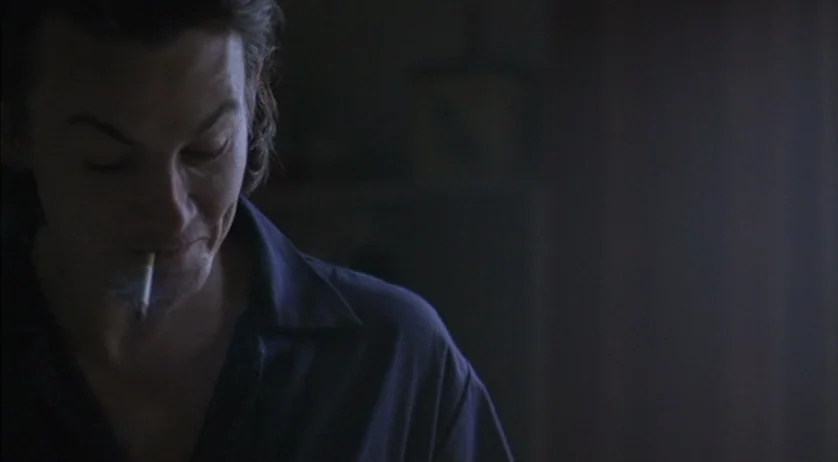 Ratcatcher (1999) movie still: medium — A dimly lit room with dark surroundings and subtle ambient shadows. A man stares downward with a lit…