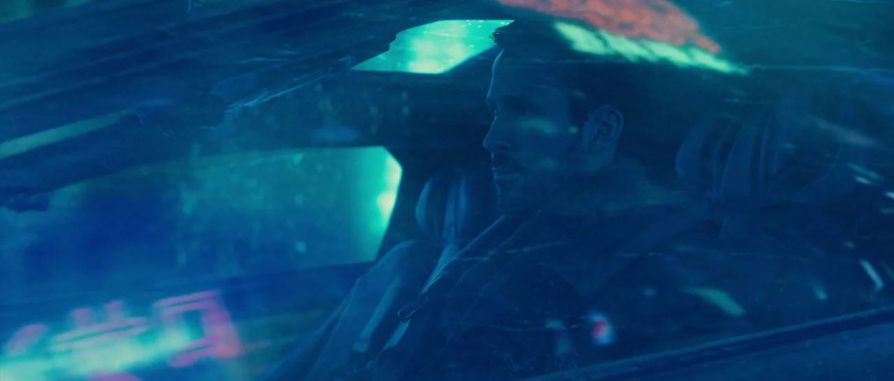 Blade Runner 2049 (2017) movie still: medium — A dark car interior at night illuminated by external neon city lights. A man sits in the driver's…