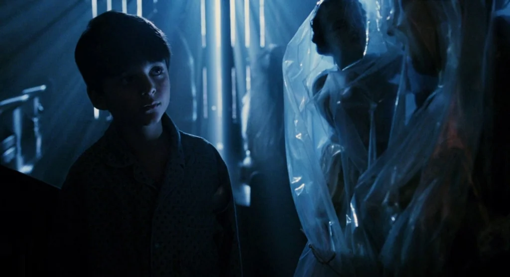 Mimic (1997) movie still: medium — A dark space features furniture draped in translucent plastic sheets. A young boy stands in the foreground…