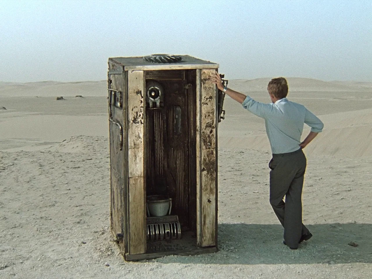 Kin-dza-dza! (1986) movie still: fullbody — A vast, barren sandy desert landscape stretches toward a pale, hazy horizon. A man stands beside an…