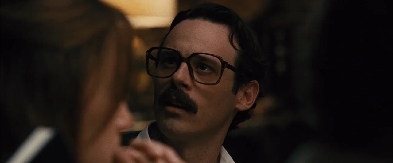 Argo (2012) movie still: closeup — A dimly lit interior room serves as the setting for a private conversation. A man with large glasses and a…
