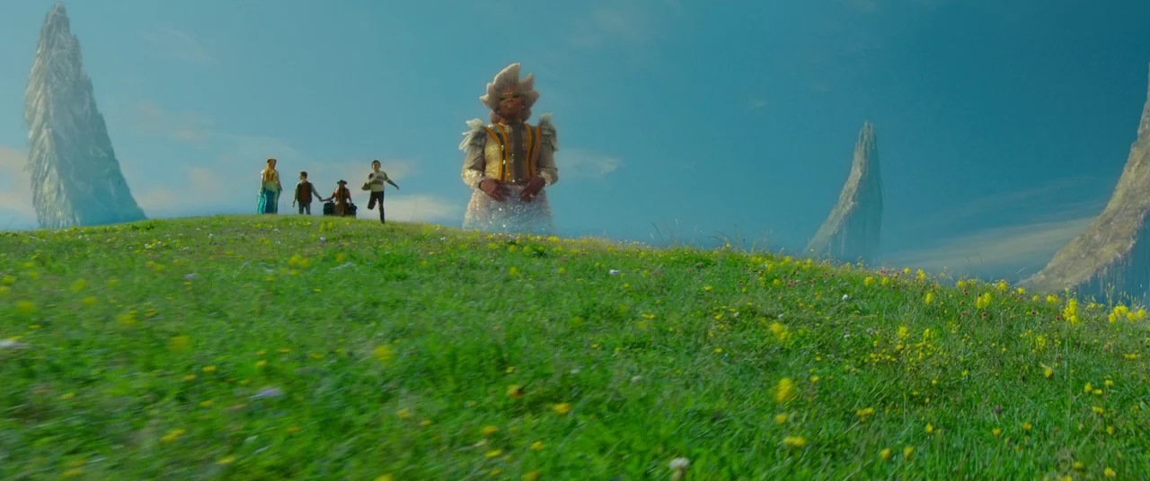 A Wrinkle In Time (2018) movie still: wide — A vast, rolling green hill is dotted with yellow flowers under a clear sky. A giant woman in shimmering…