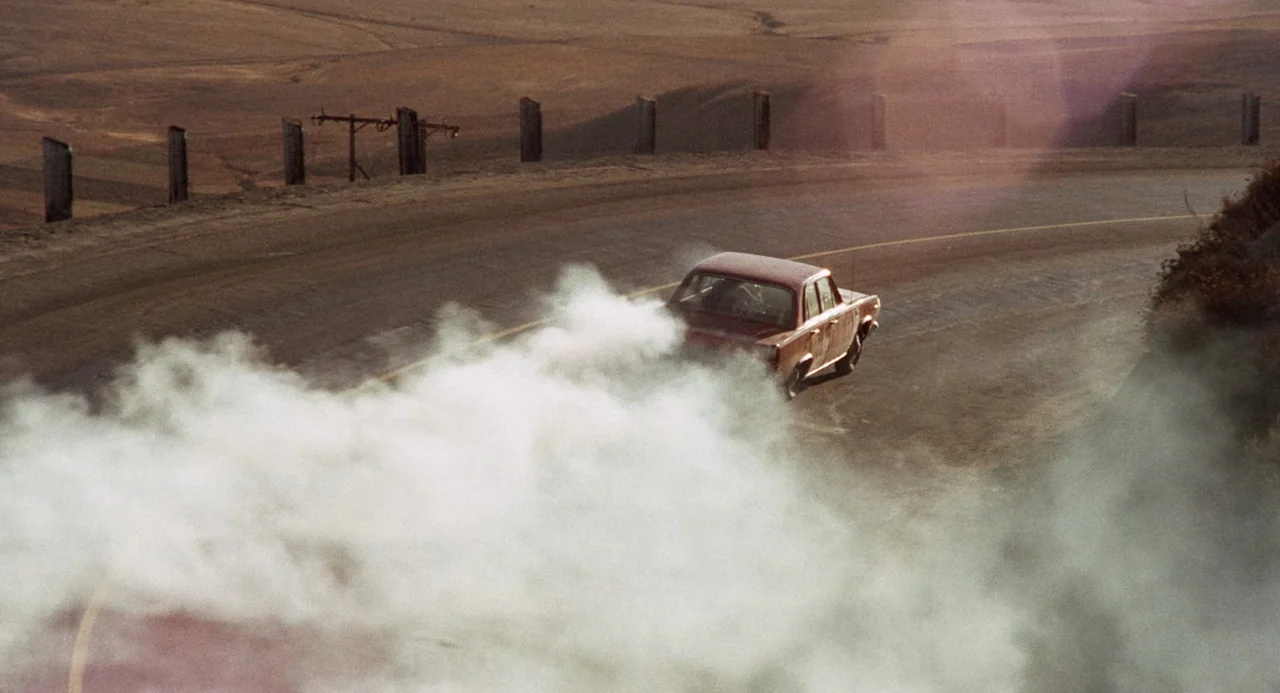 Duel (1971) movie still: wide — A dusty, sun-baked landscape features a winding road with wooden fence posts lining it. A red sedan drifts around a…
