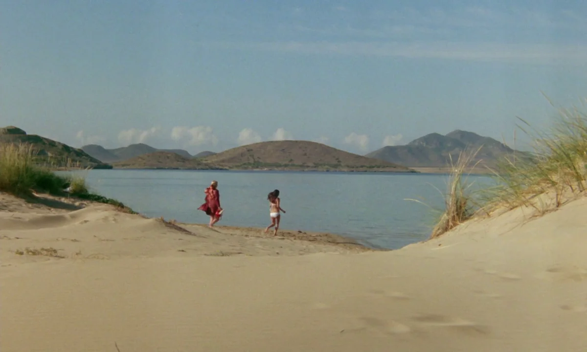 Vampyros Lesbos (1971) movie still: wide — A wide shot shows two people walking on a sandy beach by the water. Rounded hills rise above the water in…