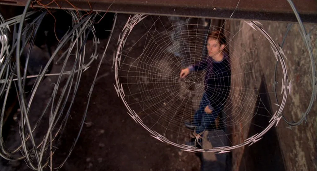 Spider-Man (2002) movie still: wide — A dark industrial space features concrete walls and tangled coils of razor wire hanging. A young man sits…