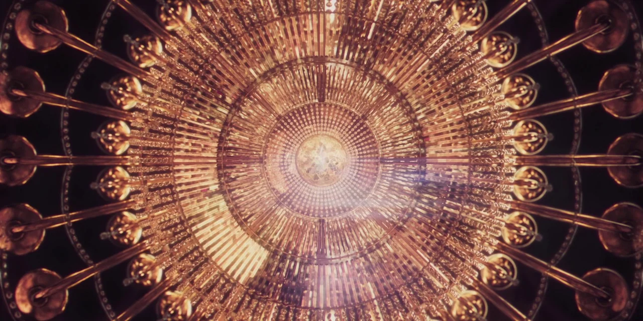 Paradise Hills (2019) movie still: closeup — A circular golden chandelier hangs from a dark ceiling in a symmetrical arrangement. The structure…