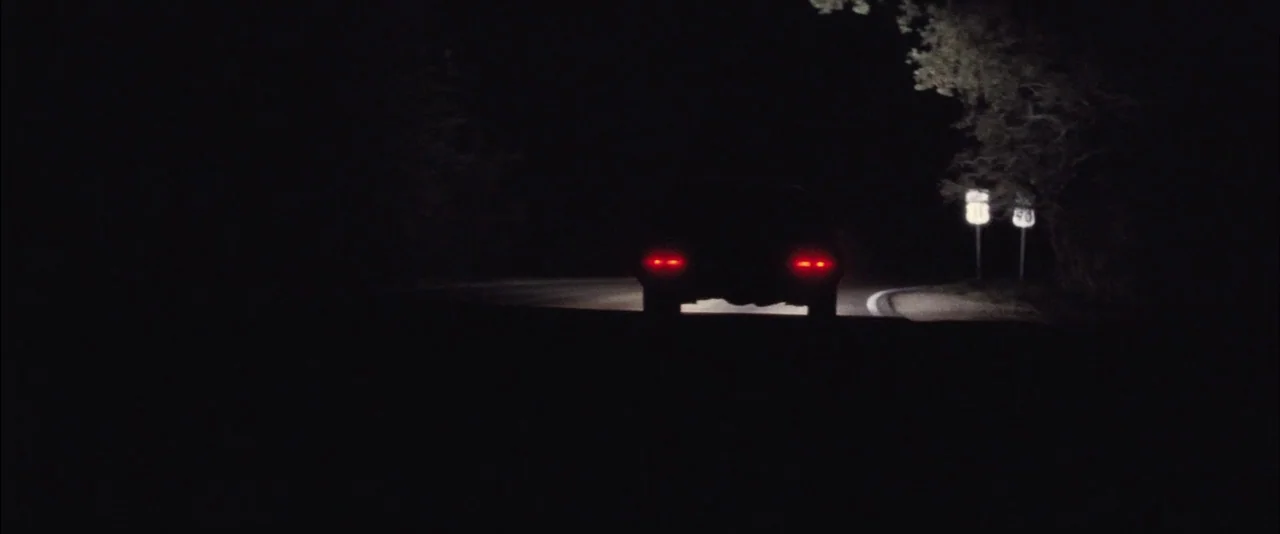 Killing Them Softly (2012) movie still: wide — A dark, empty rural road winds through a forested area at night. The taillights of a car glow red as…