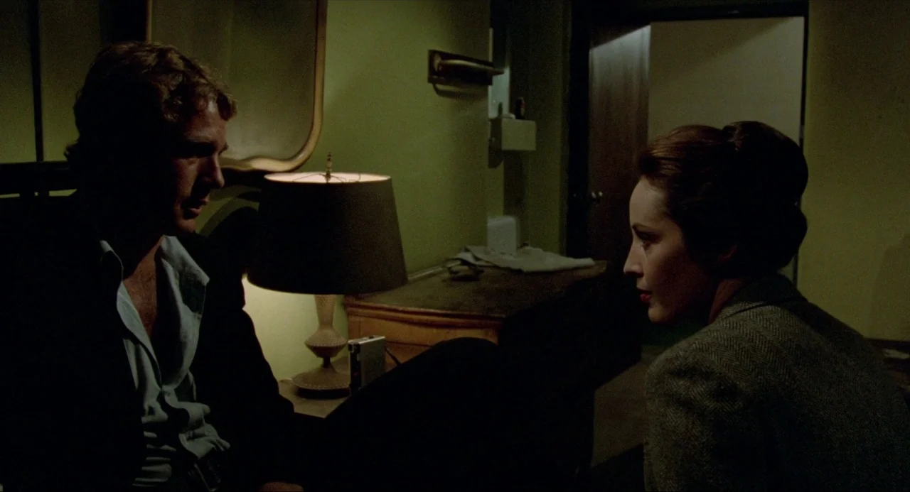 The Driver (1978) movie still: medium — A dimly lit room features a table lamp between a man and a woman. The two people sit facing each other in…
