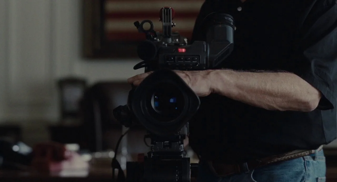 Foxcatcher (2014) movie still: medium — An interior space contains a blurry American flag hanging on a wall in back. A person holds a professional…