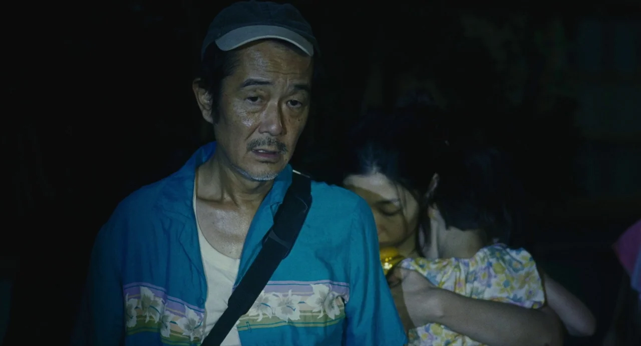 Shoplifters (2018) movie still: medium — A dark outdoor space at night with an older man and a woman. The man looks ahead while carrying the woman…