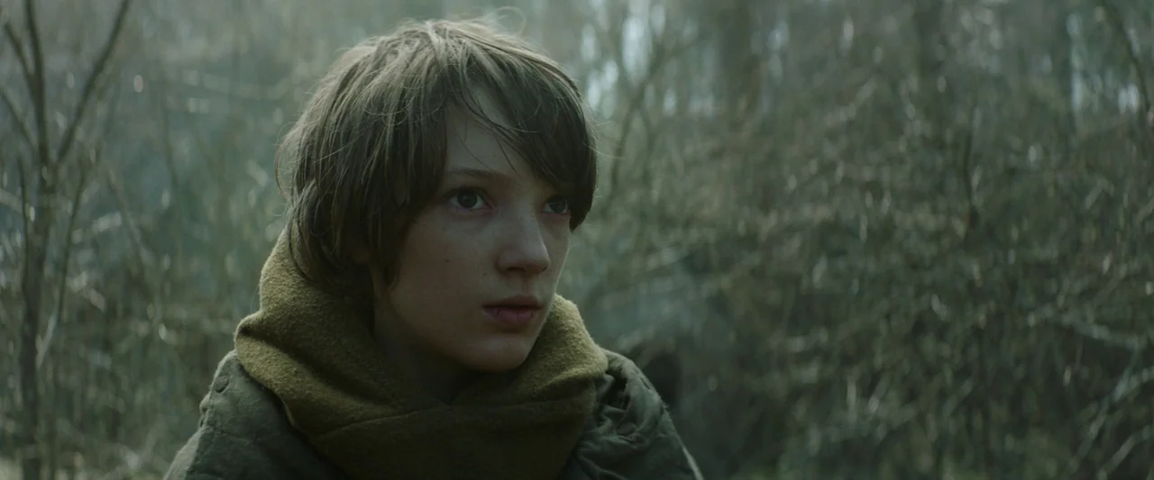 Vesper (2022) movie still: closeup — A figure stands in a dense area of leafless trees and sparse branches. The subject turns their head slightly to…