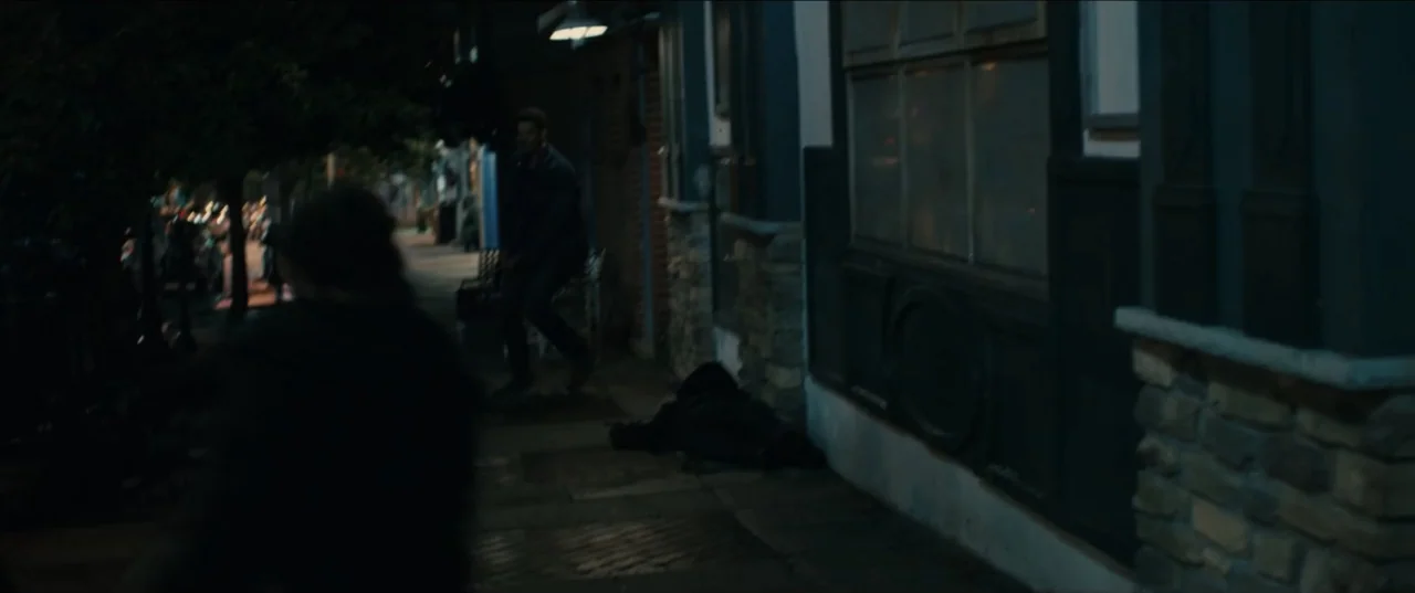 Guns Up (2025) movie still: medium — A dark urban sidewalk at night features a brick building facade and stone textures. A man in dark clothing…