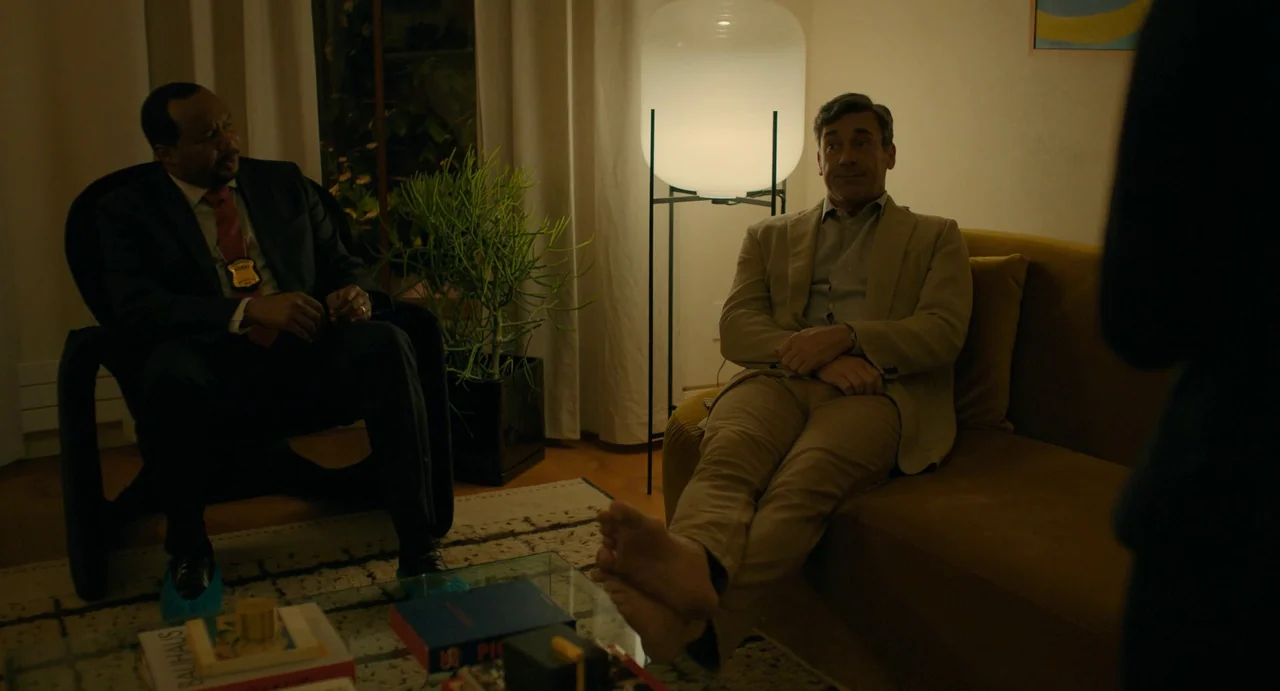 Confess, Fletch (2022) movie still: medium — A modern furnished lounge contains a mustard sofa and a glass coffee table. One man in a tan suit rests…