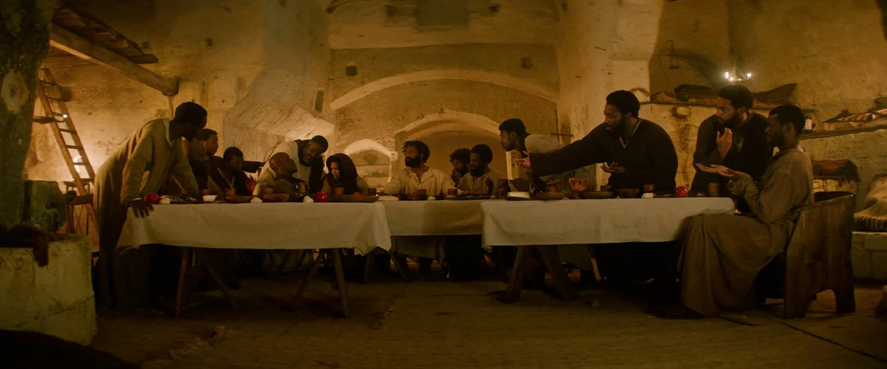 The Book of Clarence (2023) movie still: wide — A dark subterranean stone chamber with vaulted ceilings houses a long wooden table. A group of…