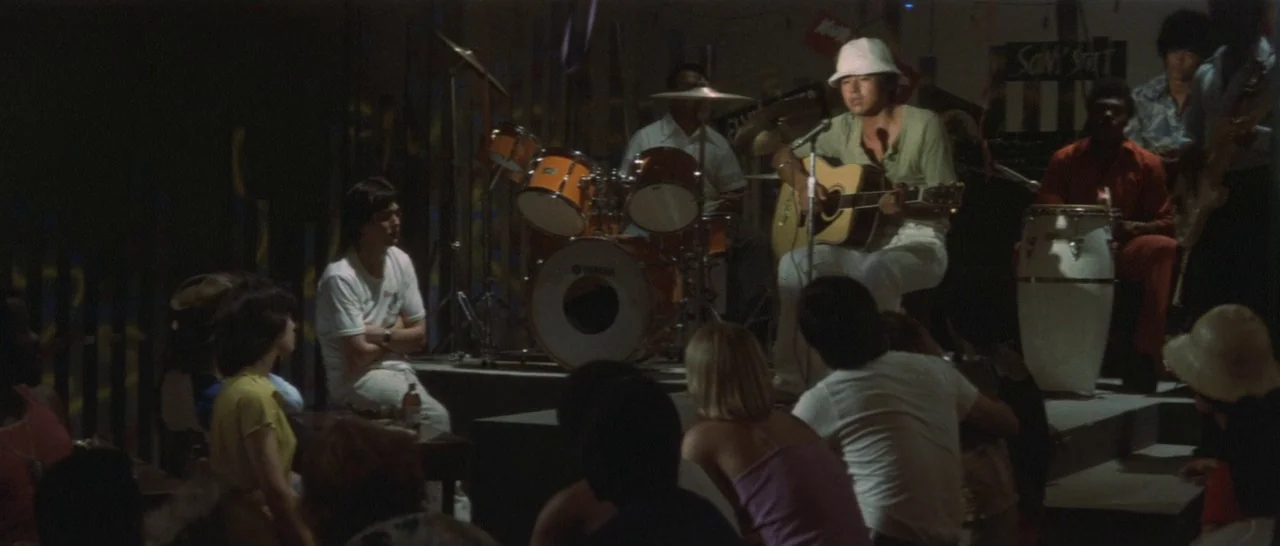 Take Me Away! (1978) movie still: wide — A dark dimly lit venue with a wooden stage holding a live band. The guitarist sits on a stool performing…