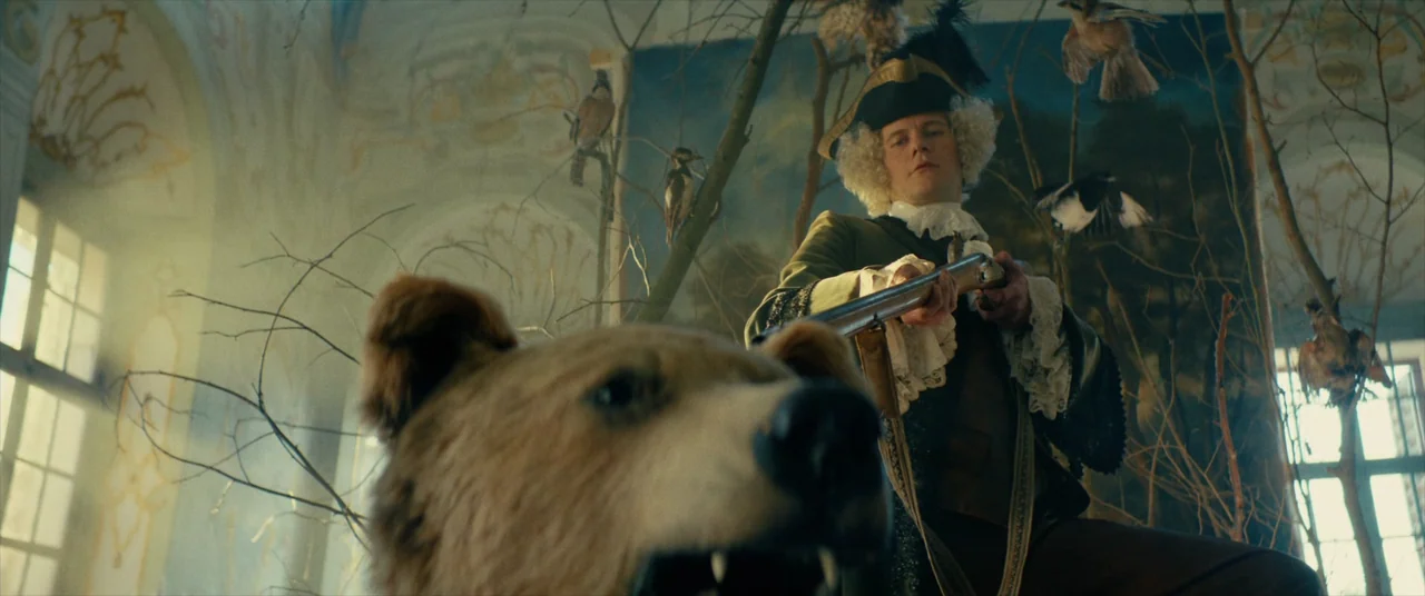 The Promised Land (2023) movie still: medium — An 18th-century style museum exhibit features taxidermy animals among bare branches. A man in a…