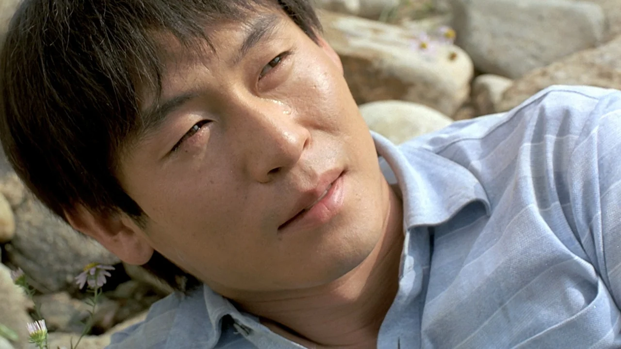 Peppermint Candy (1999) movie still: closeup — A man lies on a bed of smooth river stones and scattered wildflowers. He is positioned at an angle,…