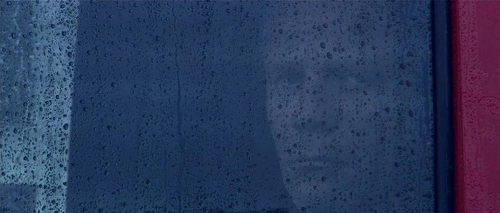 Session 9 (2001) movie still: closeup — A man's face appears as a faint reflection against a rainy dark window. Raindrops cover the surface of the…