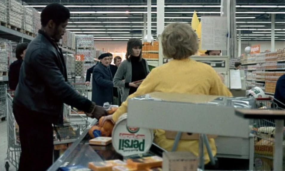Tout Va Bien (1972) movie still: medium — A busy checkout line in a 1970s supermarket with rows of inventory shelves. A man sets groceries on the…