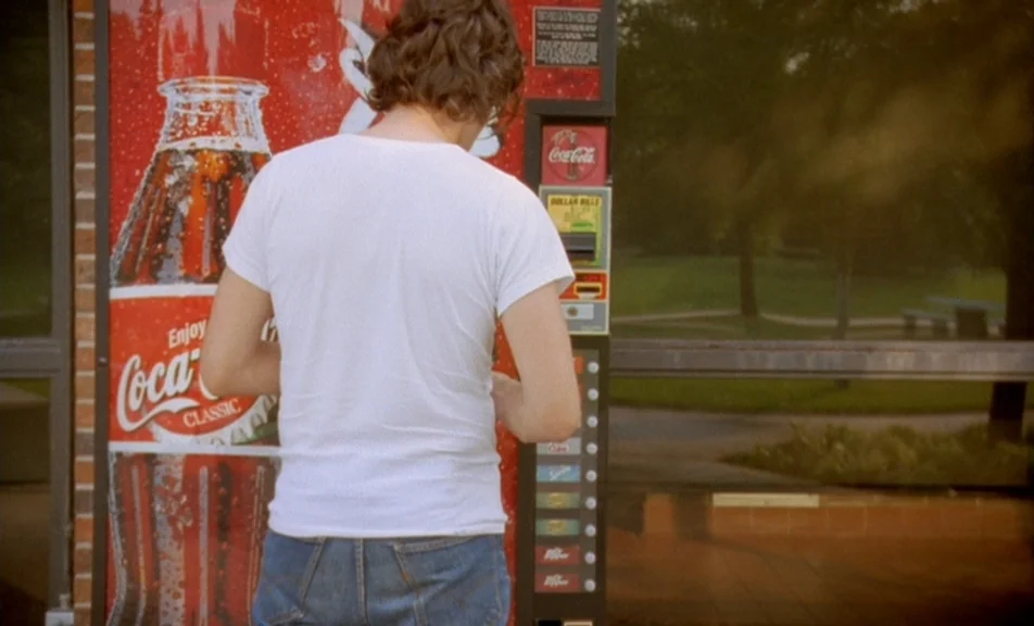 The Brown Bunny (2003) movie still: medium — A man in a white t-shirt stands before a red soda machine outdoors. The vending machine displays a…