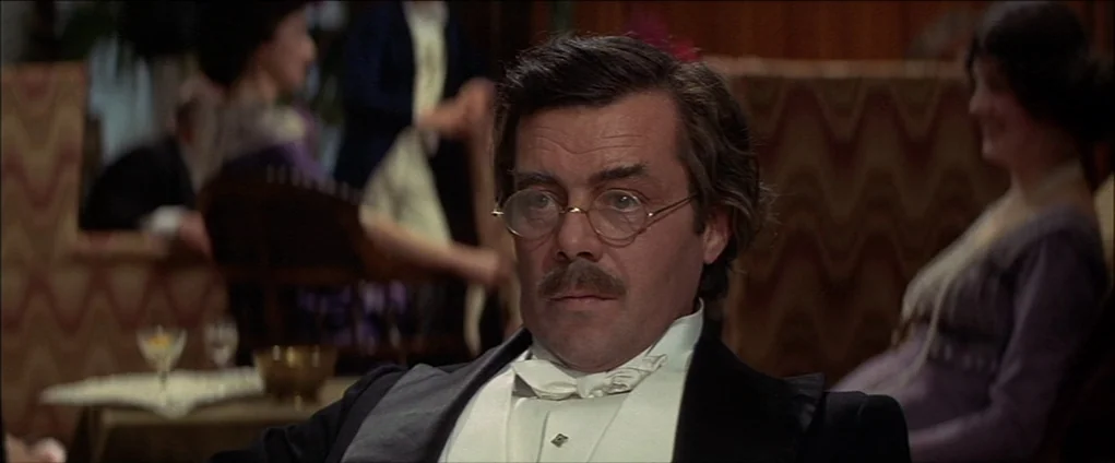 Death In Venice (1971) movie still: medium — A man sits at a table in a dimly lit, richly decorated space. Other patrons are blurred in the…