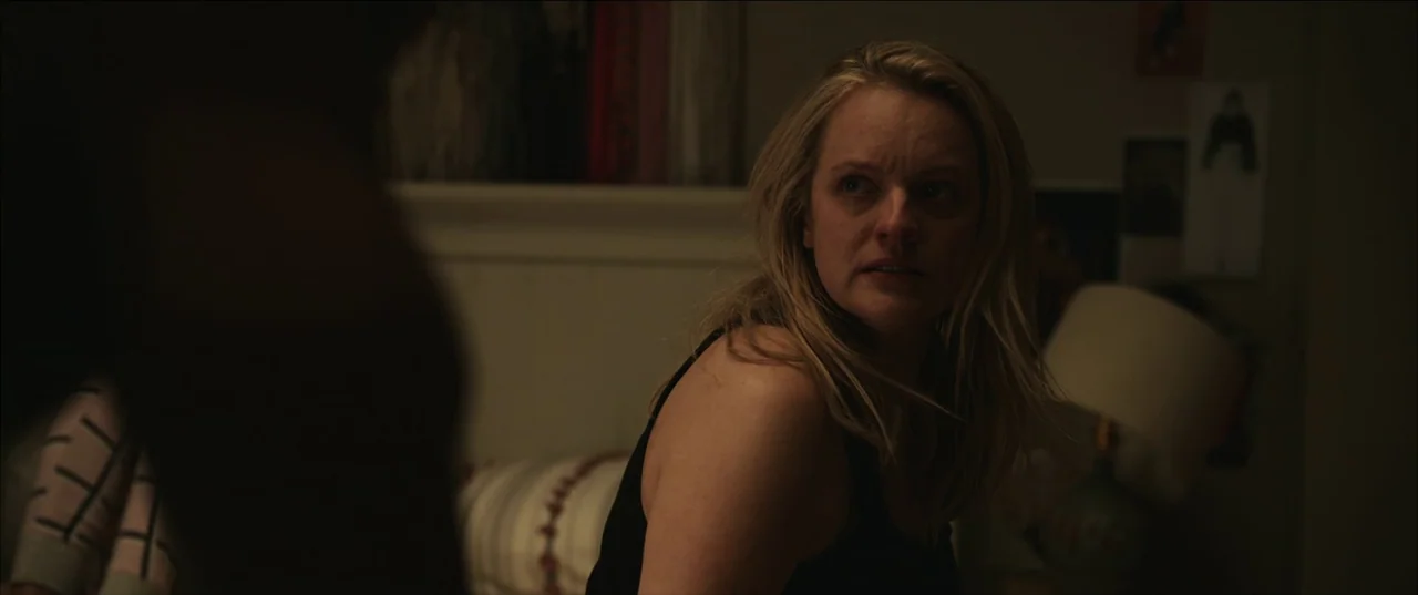 The Invisible Man (2020) (2020) movie still: medium — A dimly lit room with muted walls and a lamp in the background. A woman in a black tank top…